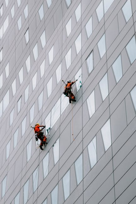 Two professional window cleaners in orange safety harnesses and helmets are suspended on a scaffold system, working on the exterior glass facade of a modern high-rise building in Ilford. The building features a pattern of large, reflective glass panels and sleek, metallic framing, with the workers using squeegees and cleaning tools to maintain the glass surfaces. The scene is well-lit with natural daylight, highlighting the cleanliness of the windows and the careful, precise cleaning process carried out by Ilford Carpet Cleaning's specialists. Although primarily focused on exterior window cleaning, the high-rise setting underscores the importance of thorough surface maintenance in commercial buildings.