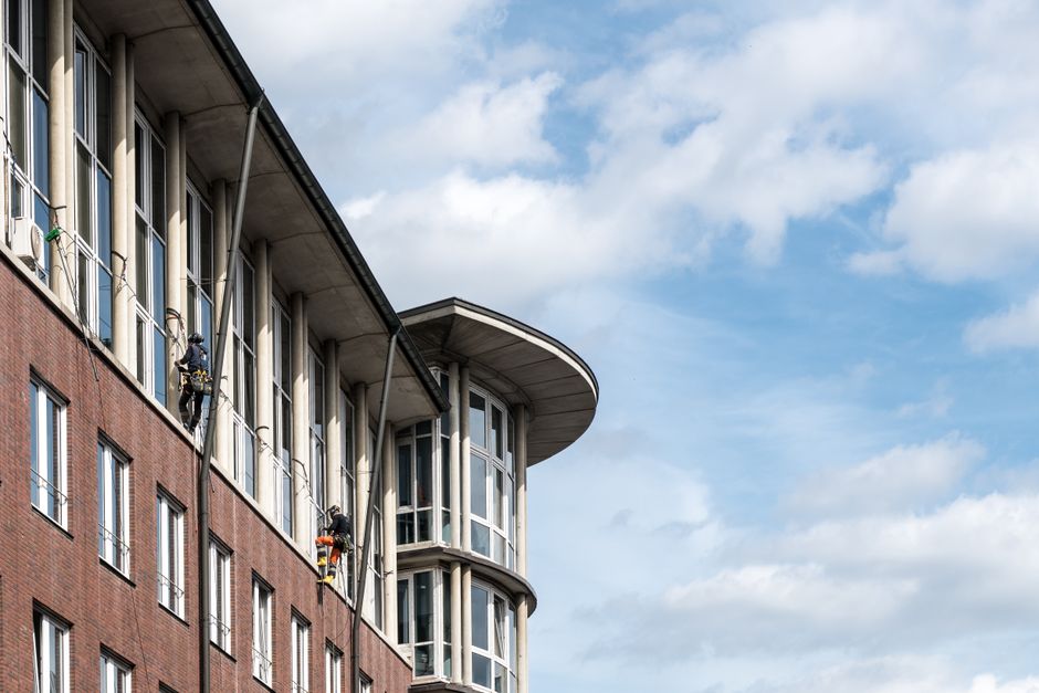 Two window cleaners are suspended on harnesses attached to the exterior of a modern high-rise building with a glass facade, performing surface cleaning on the large, reflective windows. The building features numerous glass panels framed by white structural elements, with curved balconies visible on the left side. The window cleaners are equipped with harnesses, safety gear, and cleaning tools, working amidst natural daylight that illuminates the sleek, shiny surfaces. This scene highlights the challenges of access and safety considerations for cleaning high-rise windows in residential or commercial building environments, as addressed by Ilford Carpet Cleaning in their expertise in maintaining hygienic, well-maintained spaces despite tough access issues.