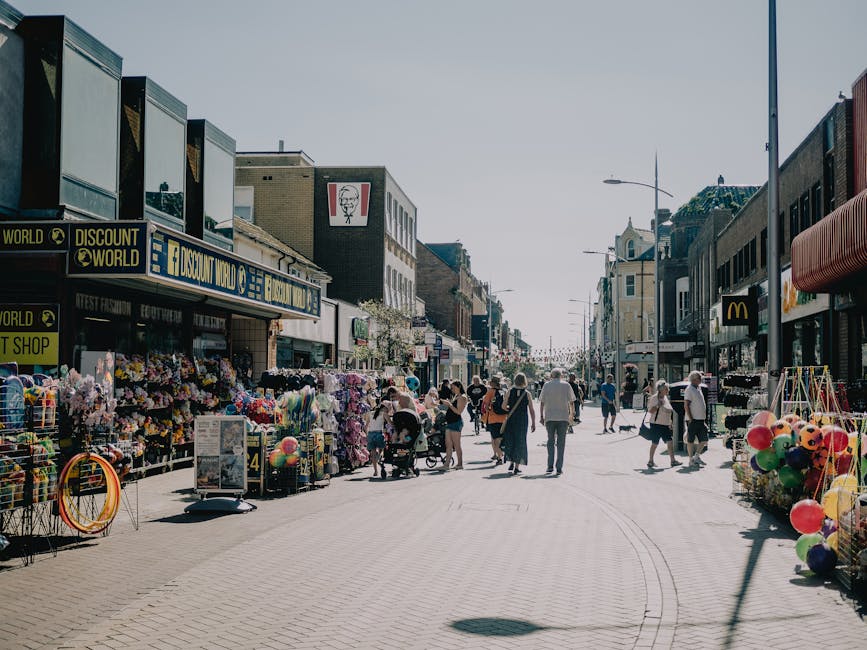 A busy street scene in Ilford during daytime, featuring various retail shops and outdoor stalls on both sides of the paved walkway. The storefronts include a discount store, a fast-food outlet, and a shop with balloons displayed outside. Pedestrians, including families, couples, and individuals, are walking along the street, some pushing strollers or carrying shopping bags. Bright daylight illuminates the scene, with clear skies overhead. The buildings are primarily made of brick, with signage and awnings in various colors, creating a lively urban atmosphere. The street appears clean and well-maintained, with no visible litter or debris, suggesting effective cleaning and maintenance by local services such as Ilford Carpet Cleaning.