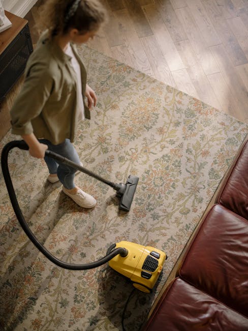A woman using a yellow cylinder vacuum cleaner with a black hose to deep clean a patterned area rug on a wooden floor in a living room. The rug features floral motifs in muted shades of orange, green, and beige. She is wearing a beige jacket, white sneakers, and dark jeans, standing near a red leather sofa. The room is well-lit with natural light, and the floor and rug appear clean and dust-free, illustrating professional surface cleaning as provided by Ilford Carpet Cleaning. The scene emphasizes hygiene and maintenance of domestic flooring surfaces.