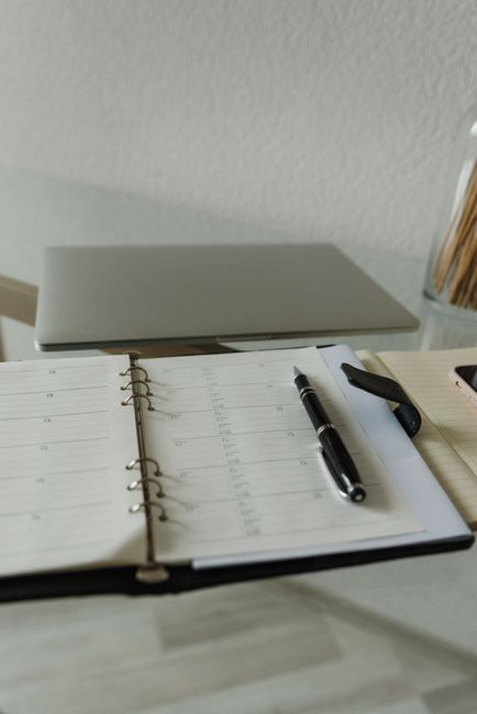 A desk surface with an open planner displaying a weekly schedule, accompanied by a black pen and a black highlighter. The desk appears clean and organized, with a closed silver laptop situated in the background. The lighting is neutral, highlighting the smooth, light-colored wooden surface of the desk. To the right, there's a small section of a container filled with various writing instruments, and the background features a plain white wall. This setting reflects a tidy workspace suitable for planning and administrative tasks, which could be part of domestic or commercial cleaning service scheduling at Ilford Carpet Cleaning, illustrating their focus on thorough surface cleaning and organisation in alignment with their booking services.
