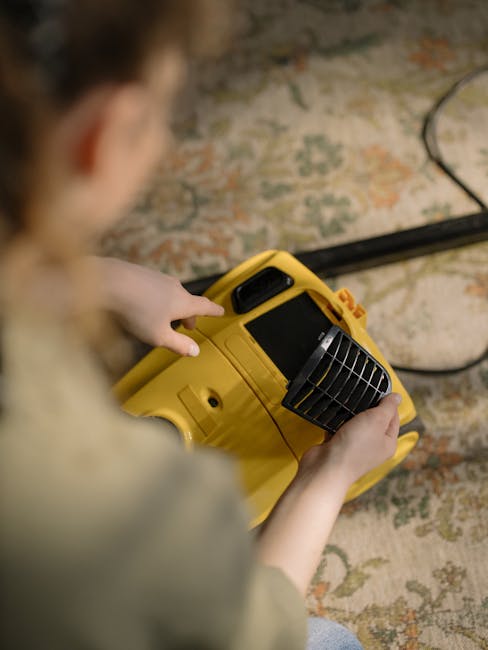 A person holding a yellow portable surface cleaner with a black vent and digital display, inspecting or preparing it for use on a patterned, beige and green carpet in a room with warm lighting. The background shows a section of the carpeted floor, suggesting a home setting. The image emphasizes the equipment used for professional deep cleaning and sanitisation, consistent with services offered by Ilford Carpet Cleaning in Barkingside and Seven Kings IG2 IG3, ILFORD.