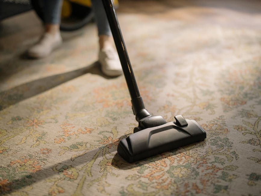 A person holding a yellow portable surface cleaner with a black vent and digital display, inspecting or preparing it for use on a patterned, beige and green carpet in a room with warm lighting. The background shows a section of the carpeted floor, suggesting a home setting. The image emphasizes the equipment used for professional deep cleaning and sanitisation, consistent with services offered by Ilford Carpet Cleaning in Barkingside and Seven Kings IG2 IG3, ILFORD.