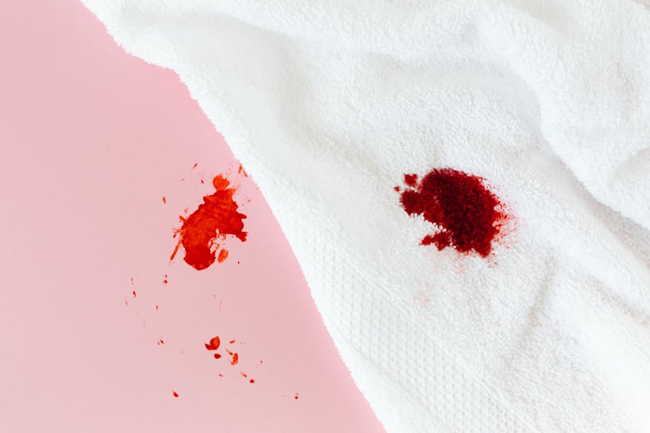 A close-up image showing two bloodstains on different surfaces; one on a light pink, slightly textured fabric and the other on a white, textured paper towel. The bloodstains are irregular in shape, with the one on the fabric appearing smeared and the one on the paper towel being more concentrated and rounded. The lighting is bright and even, highlighting the contrast between the dark red bloodstains and the pale background surfaces. This image exemplifies common household or medical spill scenarios that may require professional cleaning and stain removal, as provided by Ilford Carpet Cleaning, experts in surface cleaning and sanitisation services.