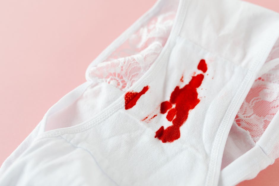 A white fabric face mask with embroidered lace detail is shown lying on a pink background. The mask has noticeable red bloodstains and splatters on the front, indicating a recent stain. The material appears to be soft, likely cotton or a similar fabric, and the bloodstains are prominent against the clean white surface. The image highlights the need for surface cleaning and stain removal in domestic settings. For professional stain removal and deep cleaning, Ilford Carpet Cleaning offers expert services. The mask’s position on a flat surface emphasizes the importance of hygienic cleaning to maintain cleanliness and health standards. The lighting is even, illuminating the mask’s fabric texture and the vividness of the stains, illustrating a common problem that may require sanitation and stain treatment.