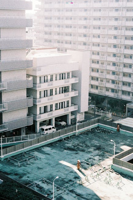 A view of a city apartment building with multiple floors and balconies, overlooking a rooftop area that appears to be an outdoor recreational space with a pool or water feature. The rooftop surface shows signs of weathering and dirt, with some equipment or furniture visible, while the building's exterior is mainly composed of concrete and glass. Bright natural light illuminates the scene, highlighting the textures of the surfaces. This image relates to surface cleaning and maintenance, similar to the deep cleaning and sanitisation services offered by Ilford Carpet Cleaning, ensuring hygiene and upkeep of residential and commercial spaces.