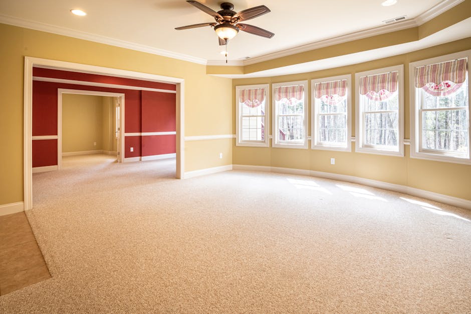 A spacious living room with light beige carpet flooring, showing a clean and well-maintained surface. The room features a large bay window with five sections, each fitted with white-framed glazing and patterned pink valances. Sunlight streams through the windows, illuminating the area. The walls are painted in a soft yellow shade, accented by a white chair rail molding running horizontally around the room. In the center of the ceiling, there is a brown ceiling fan with a light fixture. To the left, an open doorway leads to an adjoining room with a similar cream-colored carpet and a red accent wall with white trim. The room is neat, with no visible dust or stains, demonstrating effective surface cleaning in preparation for or following deep cleaning and sanitisation processes, exemplifying the quality standards offered by Ilford Carpet Cleaning.