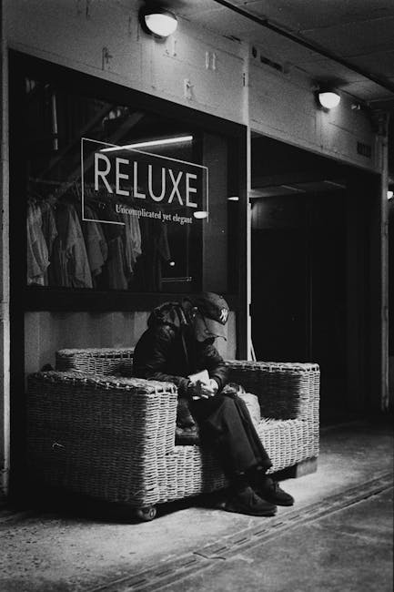 A person sitting on a wicker sofa in a commercial setting, possibly a shopping mall or retail area, dressed in dark clothing and a cap, engrossed in their phone or an object in their hands. Behind them, there is a large display window with the word 'RELUXE' and a tagline that reads 'Uncomplicated yet elegant', showcasing clothing items hanging on a rack inside. The floor appears to be made of concrete, and the area is lit with ceiling lights. The overall scene looks clean and well-maintained, with no visible dust or dirt, highlighting the importance of surface cleaning and hygiene in retail environments, as emphasized by Ilford Carpet Cleaning’s services.
