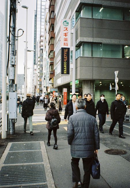 A busy urban street scene showing pedestrians walking on the sidewalk in front of modern multi-storey commercial buildings with glass windows and concrete facades in Ilford. The pavement is clean and well-maintained, with visible tactile paving and drainage grates. Some pedestrians are carrying bags or backpacks, and several are wearing face masks. The image captures a typical city environment associated with commercial activity, aligning with the context of surface cleaning and maintenance, although no cleaning tools or products are visible. Ilford Carpet Cleaning emphasizes the importance of maintaining clean surfaces in busy environments, as highlighted by the page title about permitted commercial cleaning in Redbridge, ILFORD.