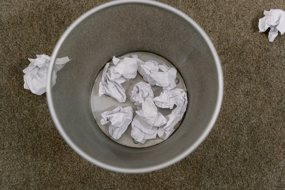 An overhead view of a cylindrical metal bin placed on a brown carpeted floor, with several crumpled white paper towels inside and a few scattered around the bin. The bin appears to be nearing capacity, and the scene suggests recent paper waste from cleaning or tidying activities. The lighting is even, highlighting the contrast between the metal surface and the textured carpet, and the environment indicates an indoor space likely used for domestic or commercial cleaning purposes, with Ilford Carpet Cleaning specializing in surface cleaning and hygiene maintenance visible in the context of waste disposal regulations after carpet cleaning.
