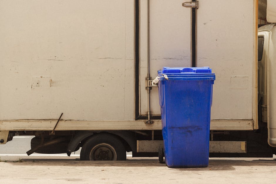 A blue waste bin is positioned next to the side of a white delivery truck with visible signs of dirt and grime. The bin is situated on a concrete surface, with the truck's lower panel and a small part of its wheel visible beneath it. The background includes the truck's white door with metal hinges and a handle, and the scene is illuminated by natural light, highlighting the cleanliness and condition of the surfaces. This setting is related to waste management practices following cleaning activities, as discussed on the page about Redbridge Council rules for waste after carpet cleaning, ILFORD, by Ilford Carpet Cleaning.
