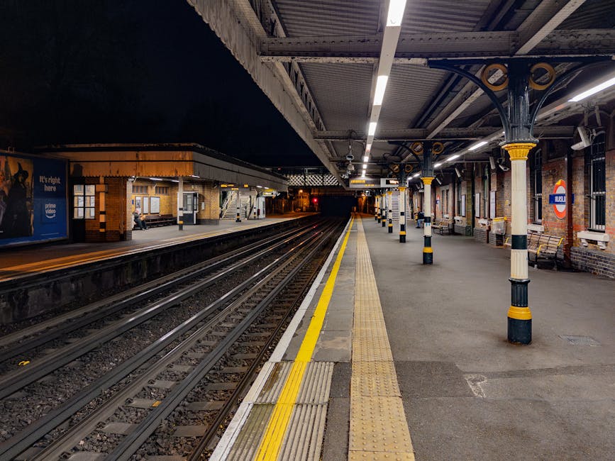 A photograph of Ilford Station at night, showing an empty platform with clean, smooth concrete surfaces. The platform features tactile paving and bright yellow safety lines along the edge, alongside evenly spaced black and yellow bollards. The station has a steel and brick structure with large windows, and the ceiling is supported by decorative columns with yellow accents. The area appears well-maintained and free of dust or debris, reflecting professional surface cleaning and upkeep by Ilford Carpet Cleaning as part of local hygiene standards for public spaces.