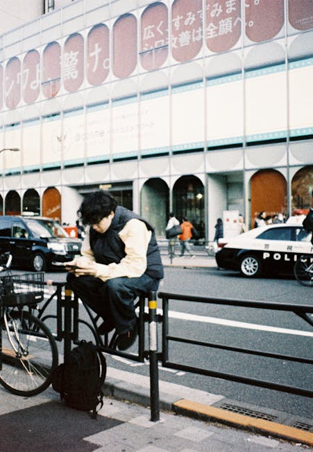 A young man dressed in a black and beige jacket seated on a black metal railing outside Ilford Station, with a black backpack resting on the ground beside him. He is focused on his smartphone, with pedestrians walking nearby and a police car parked on the street. The modern station building behind features a glass facade with signage in Japanese characters and large arched windows. The scene captures a typical urban setting emphasizing cleanliness and order associated with professional cleaning and maintenance services, reflecting the importance of surface cleanliness in busy public areas. Image is well-lit, highlighting the urban environment and the station's architectural details, consistent with a commercial or public space recently maintained for hygiene and safety, as promoted by Ilford Carpet Cleaning.