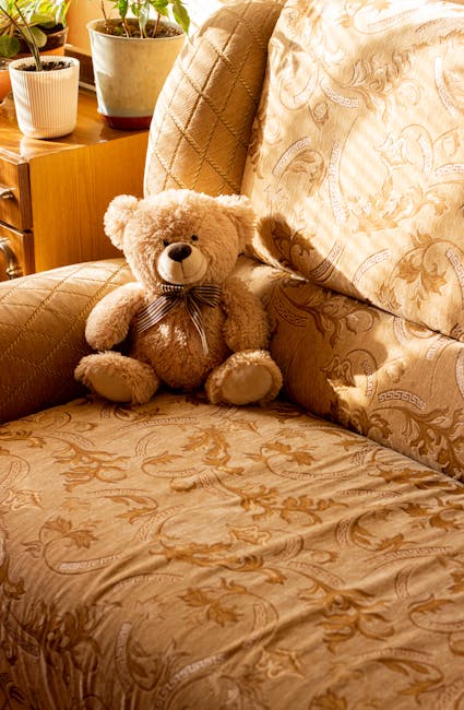 A close-up view of a beige upholstered sofa with a floral and damask pattern, featuring a soft, plush teddy bear with a striped ribbon around its neck sitting on the seat cushion. The sofa is situated in a well-lit living room with a wooden side table behind it, decorated with potted plants in cream and terracotta containers. The cushions and upholstery appear clean and well-maintained, with no visible dust or stains, highlighting professional surface cleaning and maintenance associated with domestic cleaning services provided by Ilford Carpet Cleaning for upholstery in Ilford's Valentines Park area.