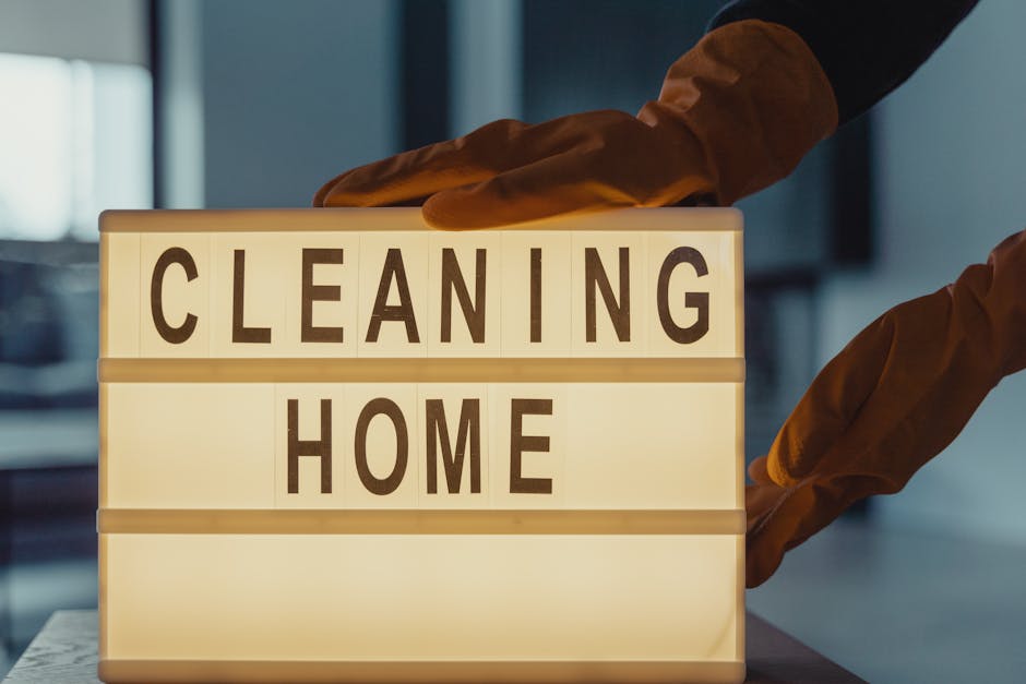 A person wearing orange rubber gloves is adjusting a backlit sign displaying the words 'CLEANING HOME' in large black letters on a white background, placed on a table in a well-lit room. The room features a modern kitchen with a dark countertop, metallic appliances, and a window allowing natural light, emphasizing clean and hygienic surface cleaning suitable for end of tenancy requirements. This visual relates to professional domestic cleaning services provided by Ilford Carpet Cleaning, highlighting surface disinfection and deep cleaning for residential spaces.