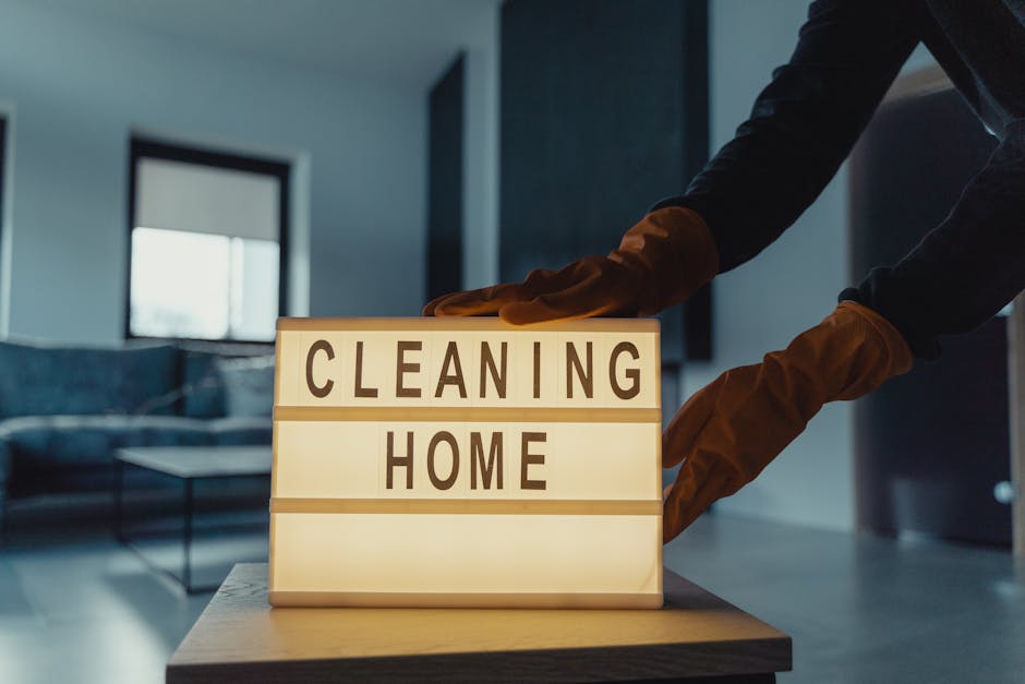 A person wearing black clothing and brown cleaning gloves is seen placing a small illuminated sign on a light-colored wooden table in a modern living room. The sign displays the words 'CLEANING HOME' in bold letters on separate lines. The room features a dark sofa with a blue cushion, a glass coffee table, and a large window allowing natural light to fill the space. The floor appears to be light-colored wood, and the overall atmosphere is clean and tidy, reflecting professional surface cleaning or deep cleaning activities as part of residential hygiene maintenance. Natural daylight enhances the room's brightness, and the scene emphasizes the importance of domestic cleaning services, such as those offered by Ilford Carpet Cleaning, in maintaining a hygienic living environment.