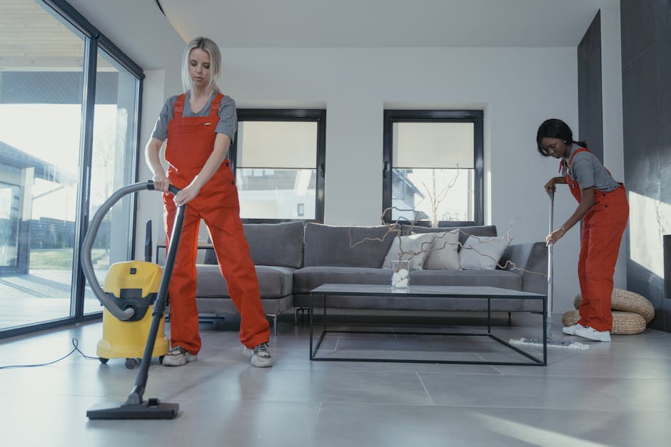 A person wearing orange rubber gloves is adjusting a backlit sign displaying the words 'CLEANING HOME' in large black letters on a white background, placed on a table in a well-lit room. The room features a modern kitchen with a dark countertop, metallic appliances, and a window allowing natural light, emphasizing clean and hygienic surface cleaning suitable for end of tenancy requirements. This visual relates to professional domestic cleaning services provided by Ilford Carpet Cleaning, highlighting surface disinfection and deep cleaning for residential spaces.